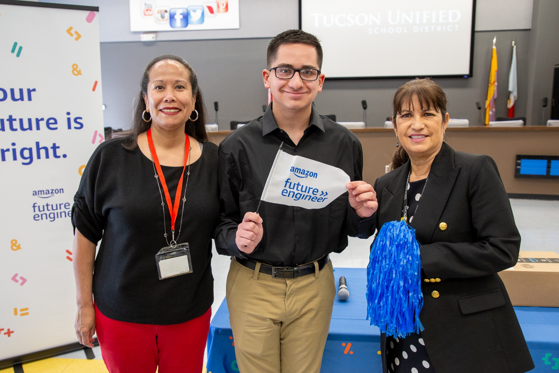 ߲о High senior Anthony Talavera smiles and poses with an Amazon Future Engineer flag and two TUSD staff members
